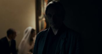 Movie still from “Certified Copy” (2010), directed by Abbas Kiarostami – A man standing in a dark room with a woman in the background; Close Up shot, Over the shoulder angle