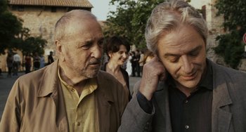 Movie still from “Certified Copy” (2010), directed by Abbas Kiarostami – Two older men standing next to each other on the street; Close Up shot, Over the shoulder angle