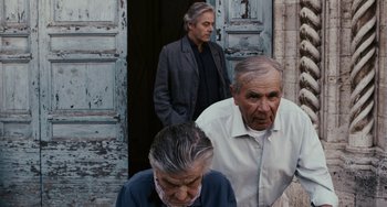 Movie still from “Certified Copy” (2010), directed by Abbas Kiarostami – A group of people standing next to each other near a building; Medium shot, Over the shoulder angle
