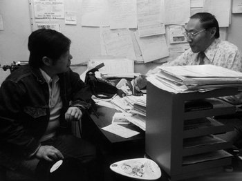 Movie still from “Chan Is Missing” (1982), directed by Wayne Wang – Two people sitting at a desk in front of a stack of papers; Medium shot, High angle