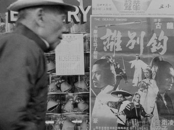 Movie still from “Chan Is Missing” (1982), directed by Wayne Wang – An old man in a hat walking past a poster of a movie; Close Up shot, Over the shoulder angle