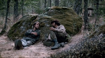 Movie still from “Che: Part Two” (2008), directed by Steven Soderbergh – Two men sitting on the ground in the woods; Wide shot, High angle