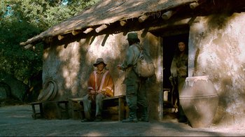 Movie still from “Che: Part Two” (2008), directed by Steven Soderbergh – A man sitting on a bench in front of a building; Wide shot, Over the shoulder angle