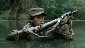 Movie still from “Che: Part Two” (2008), directed by Steven Soderbergh – A man holding a rifle in a body of water; Medium shot, High angle