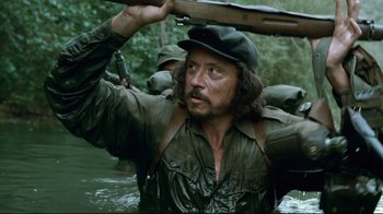 Movie still from “Che: Part Two” (2008), directed by Steven Soderbergh – A man with a rifle in a body of water; Medium shot, Over the shoulder angle