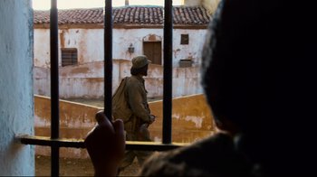 Movie still from “Che: Part Two” (2008), directed by Steven Soderbergh – A man walking through a jail cell through a fence; Wide shot, Over the shoulder angle