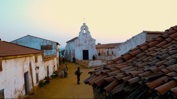 Movie still from “Che: Part Two” (2008), directed by Steven Soderbergh – A group of people standing on a dirt road near a building; Extreme Wide shot, Low angle