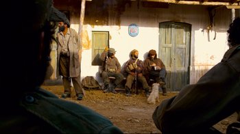 Movie still from “Che: Part Two” (2008), directed by Steven Soderbergh – A group of men sitting on a bench in front of a building; Wide shot, Over the shoulder angle