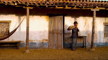 Movie still from “Che: Part Two” (2008), directed by Steven Soderbergh – A man standing in front of a building holding a piece of wood; Wide shot, Low angle