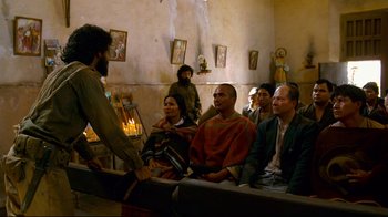 Movie still from “Che: Part Two” (2008), directed by Steven Soderbergh – A group of people sitting in a room with a priest; Wide shot, Over the shoulder angle
