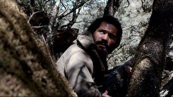 Movie still from “Che: Part Two” (2008), directed by Steven Soderbergh – A man with a gun in the woods looking at the camera; Medium shot, Low angle