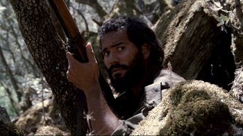 Movie still from “Che: Part Two” (2008), directed by Steven Soderbergh – A man with a beard holding a rifle in a tree; Close Up shot, Over the shoulder angle