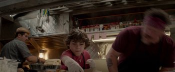 Movie still from “Chef” (2014), directed by Jon Favreau – A young boy in a red shirt and white gloves is in a kitchen; Medium shot, Over the shoulder angle