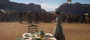 Movie still from “Cheyenne Autumn” (1964), directed by John Ford – A woman standing in front of a table with food on top of it; Wide shot, Over the shoulder angle