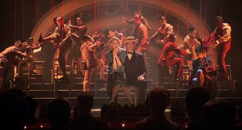 Movie still from “Chicago” (2002), directed by Rob Marshall – A man and a woman sitting on a stage with a crowd of people; Wide shot, Overhead angle