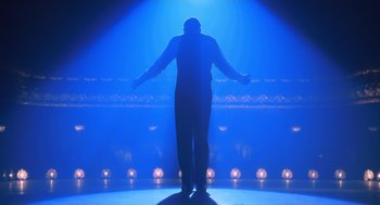 Movie still from “Chicago” (2002), directed by Rob Marshall – A man standing on a stage in front of a blue light; Wide shot, Low angle