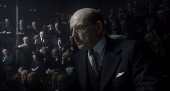 Movie still from “Darkest Hour” (2017), directed by Joe Wright – A man in a suit and tie in front of a group of people; Medium shot, Over the shoulder angle