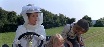 Movie still from “Chitty Chitty Bang Bang” (1968), directed by Ken Hughes – A woman driving a car while two other people sit on the back of the car; Medium shot, Over the shoulder angle