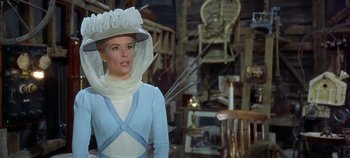 Movie still from “Chitty Chitty Bang Bang” (1968), directed by Ken Hughes – A woman wearing a white hat standing next to a chair; Medium shot, Over the shoulder angle