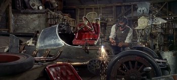 Movie still from “Chitty Chitty Bang Bang” (1968), directed by Ken Hughes – A man working on an antique car in a garage; Wide shot, Low angle