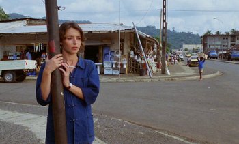 Movie still from “Chocolat” (1988), directed by Claire Denis – A woman standing next to a pole on the side of the road; Medium shot, Low angle