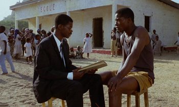Movie still from “Chocolat” (1988), directed by Claire Denis – Two men are sitting on stools and one of them is reading a book; Medium shot, Low angle