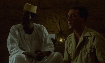 Movie still from “Chocolat” (1988), directed by Claire Denis – Two men are sitting in a room and one of them is holding a remote control; Medium shot, Over the shoulder angle