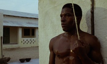 Movie still from “Chocolat” (1988), directed by Claire Denis – A shirtless man holding a stick in front of a building; Close Up shot, Low angle