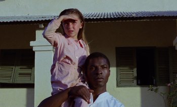 Movie still from “Chocolat” (1988), directed by Claire Denis – A young girl sitting on the shoulders of an older man; Medium shot, Low angle