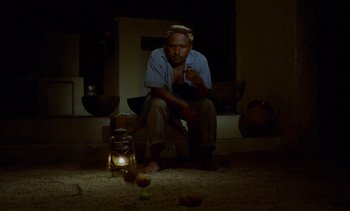 Movie still from “Chocolat” (1988), directed by Claire Denis – A man sitting on the floor in front of a lantern; Wide shot, Low angle
