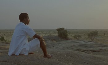 Movie still from “Chocolat” (1988), directed by Claire Denis – A man sitting on top of a rock in the desert; Medium shot, Low angle