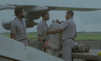 Movie still from “Chocolat” (1988), directed by Claire Denis – A group of men standing next to each other near an airplane; Wide shot, High angle