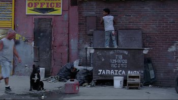 Movie still from “Chop Shop” (2007), directed by Ramin Bahrani – A man standing on top of a dumpster next to a building; Wide shot, High angle