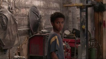 Movie still from “Chop Shop” (2007), directed by Ramin Bahrani – A young boy standing next to a pile of junk in a garage; Medium shot, Over the shoulder angle
