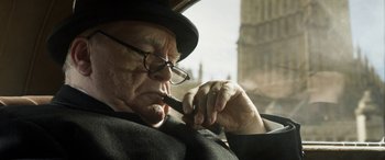 Movie still from “Churchill” (2017), directed by Jonathan Teplitzky – An older man in a hat and glasses smoking a pipe; Close Up shot, Low angle