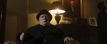 Movie still from “Churchill” (2017), directed by Jonathan Teplitzky – An older man in a hat and a coat sitting in front of a lamp; Medium shot, Low angle
