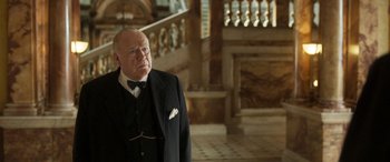 Movie still from “Churchill” (2017), directed by Jonathan Teplitzky – An older man in a suit and bow tie in front of a staircase; Medium shot, Over the shoulder angle