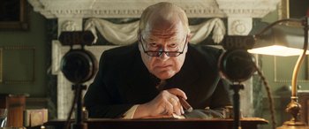 Movie still from “Churchill” (2017), directed by Jonathan Teplitzky – An older man in a black suit and glasses is leaning on a desk; Close Up shot, Low angle