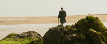 Movie still from “Churchill” (2017), directed by Jonathan Teplitzky – An old man in a black coat and hat walking on the beach; Wide shot, Low angle