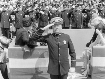 Movie still from “City Lights” (1931), directed by Charles Chaplin – An officer saluting in front of a crowd of people; Medium shot, High angle