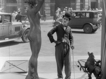 Movie still from “City Lights” (1931), directed by Charles Chaplin – An old black and white photo of a man in a suit standing next to a statue; Wide shot, Low angle