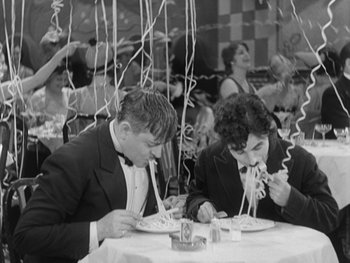Movie still from “City Lights” (1931), directed by Charles Chaplin – A black - and - white photo of two men eating at a party; Medium shot, High angle