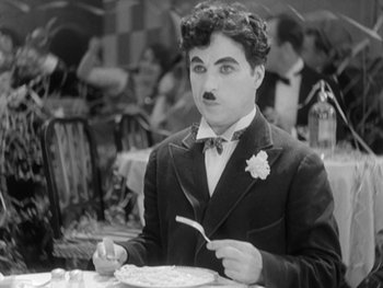 Movie still from “City Lights” (1931), directed by Charles Chaplin – A man in a suit holding a fork in his mouth; Medium shot, High angle