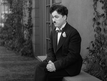 Movie still from “City Lights” (1931), directed by Charles Chaplin – A man sitting on a bench wearing a suit and tie; Medium shot, High angle