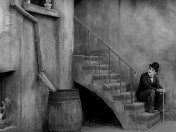 Movie still from “City Lights” (1931), directed by Charles Chaplin – A person sitting on a set of stairs wearing a mask; Wide shot, High angle