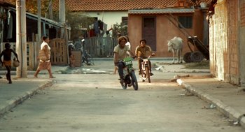 Movie still from “City of God” (2002), directed by Kátia Lund – Two people on mopeds and bicycles riding down a street; Wide shot, Low angle