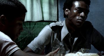 Movie still from “City of God” (2002), directed by Kátia Lund – A man sitting at a table with a glass of water in front of him; Close Up shot, Over the shoulder angle