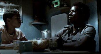 Movie still from “City of God” (2002), directed by Kátia Lund – A man sitting at a table with a basket of food; Medium shot, Over the shoulder angle