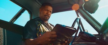 Movie still from “Day Shift” (2022), directed by J.J. Perry – A man sitting in the driver's seat of a car reading a book; Close Up shot, Low angle