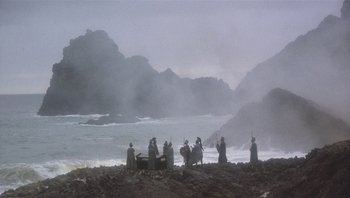 Movie still from “Clash of the Titans” (1981), directed by Desmond Davis – A group of people standing on top of a rocky beach; Extreme Wide shot, Low angle